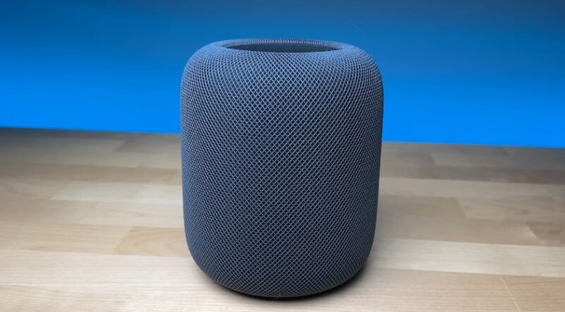 A smart, gray speaker sits on a light wooden surface