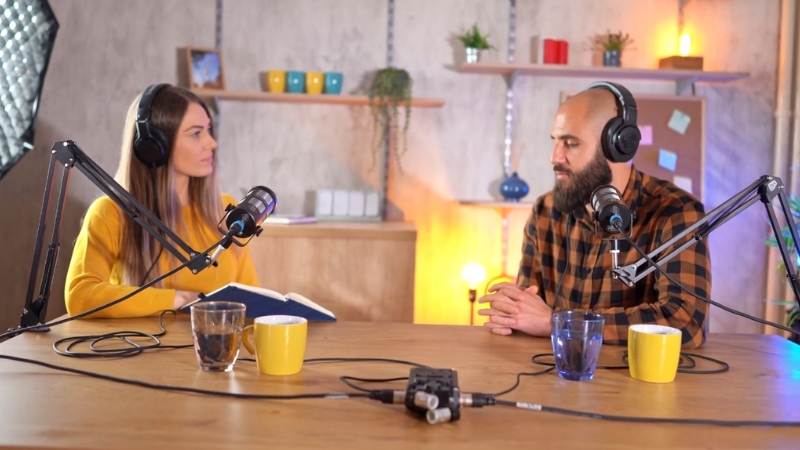 Two podcast hosts sit across from each other with mics and headphones