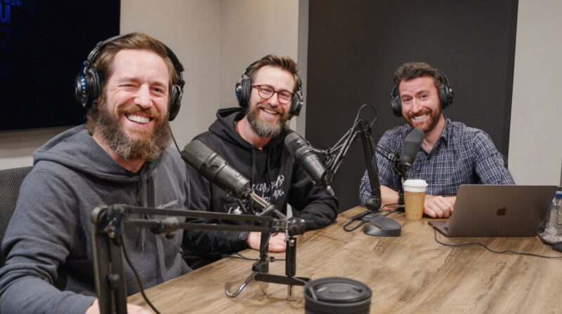 Three smiling podcasters wearing headphones and sitting at a table with microphones, coffee, and a laptop, recording an episode together