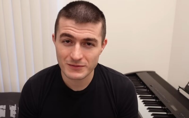 Lex Fridman sits in front of a piano, looking at camera