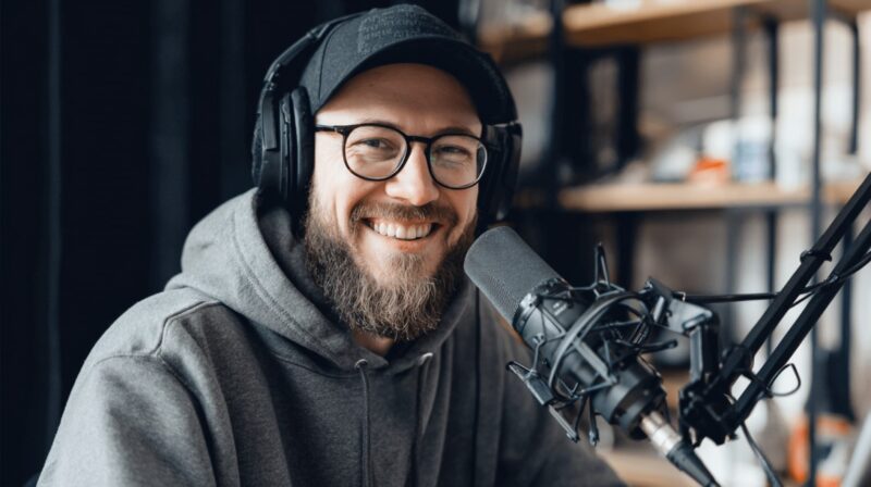 Smiling bearded man in glasses and a hoodie recording a podcast with headphones and a microphone in front of him