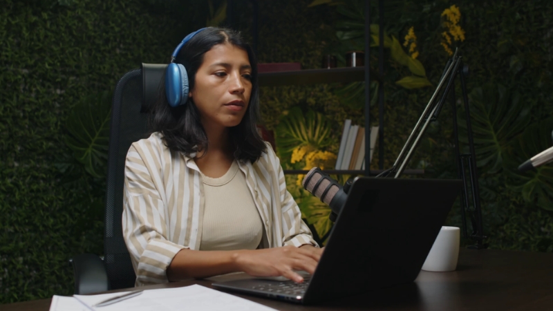 Woman with headphones working on a laptop with a microphone on the desk for a podcast hosting platform checklist