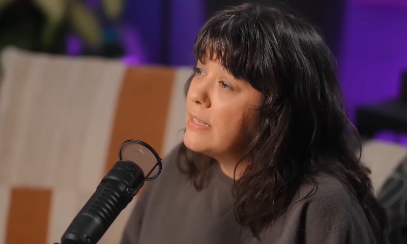 Brit Prawat speaking into a professional black studio microphone with soft lighting and a purple background accent