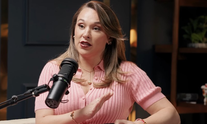 Ashley Flowers in a pink shirt speaks into a microphone during a podcast