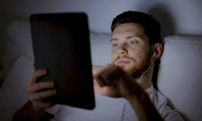 A man lies in bed in low light, using a tablet. He wears earphones and appears focused