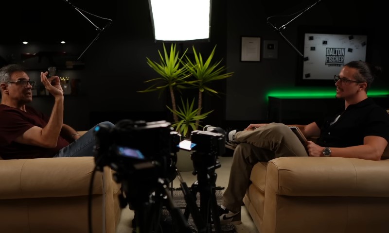 Two people are seated on beige armchairs in a dimly lit studio, engaged in an interview