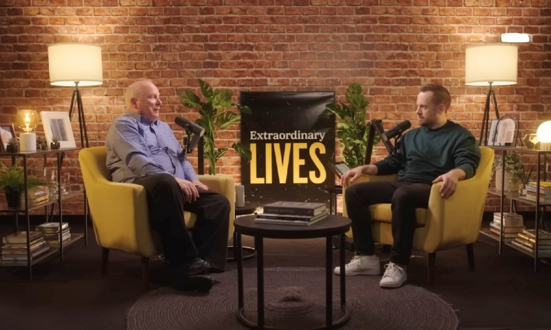 Two men sit in yellow chairs, conversing on a podcast set with a "Extraordinary Lives" banner