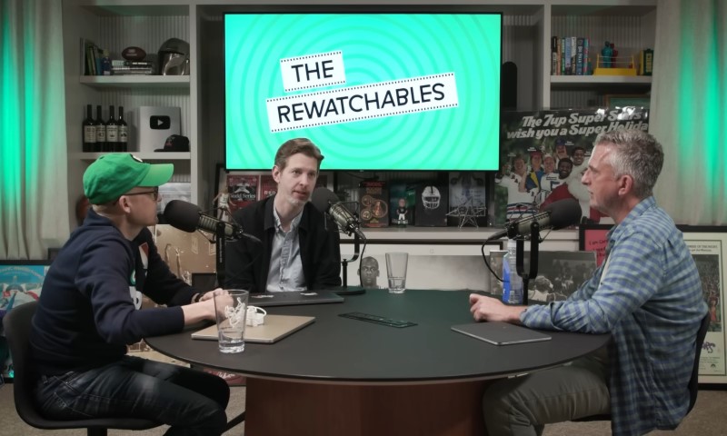 Three men are sitting around a table with microphones, recording a podcast episode. A screen behind them displays "The Rewatchables."